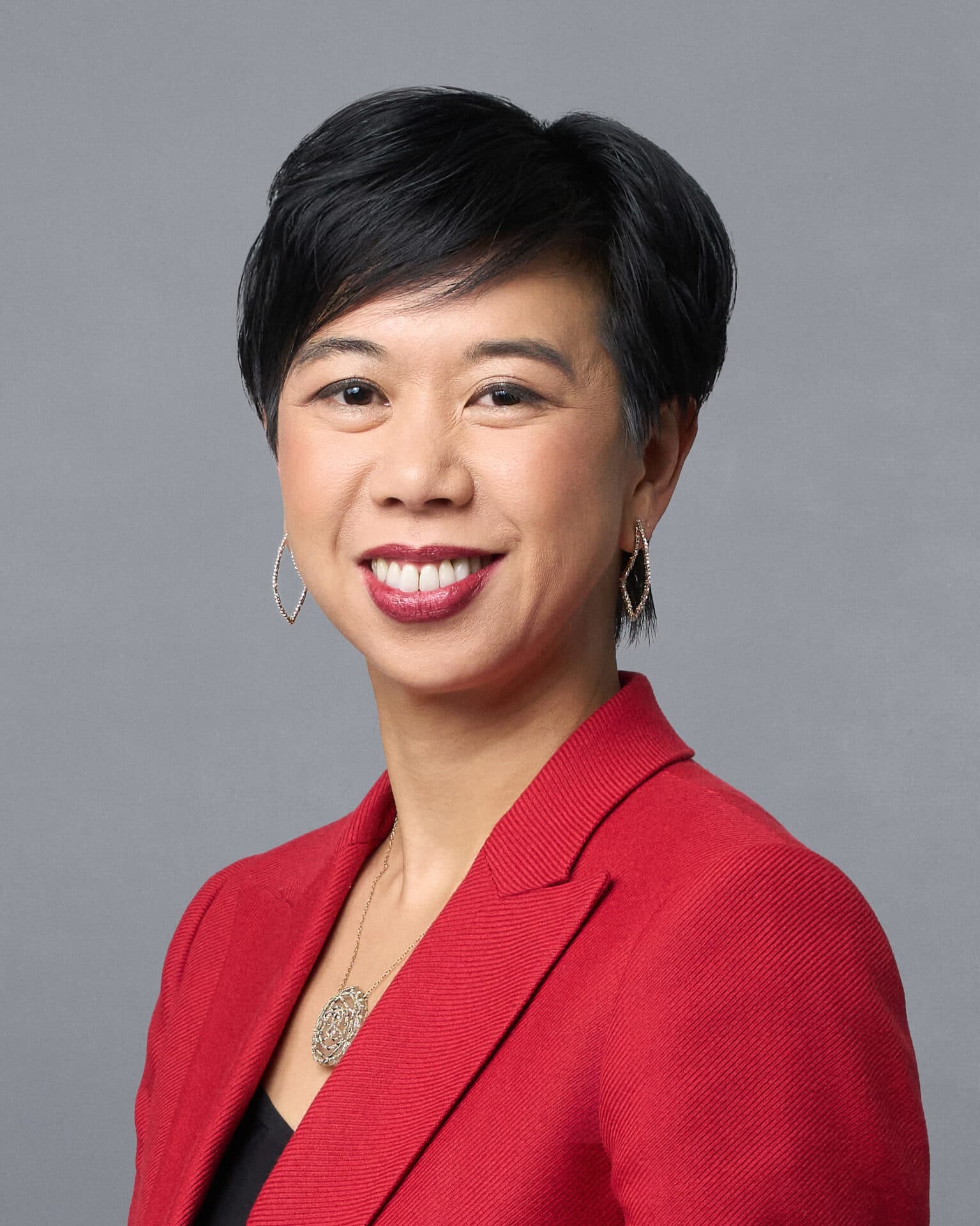 Smiling woman with short black hair wearing a red blazer and silver jewelry, gray background.