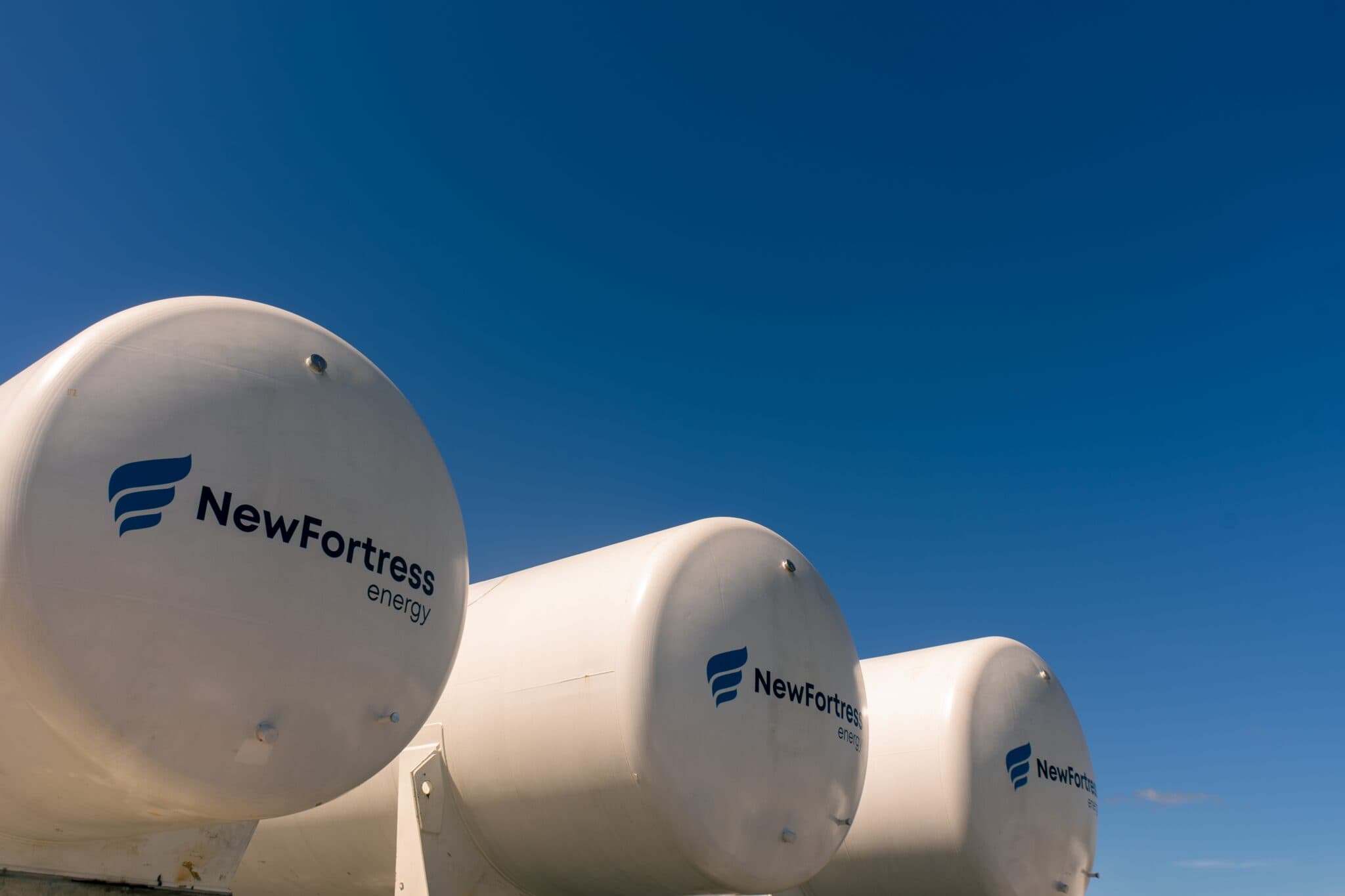 Three large white New Fortress Energy storage tanks against a clear, bright blue sky.